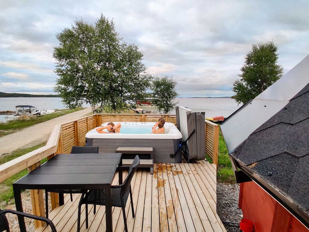 Outdoor hot tub on a terrace overlooking a lake and forest in summer light.